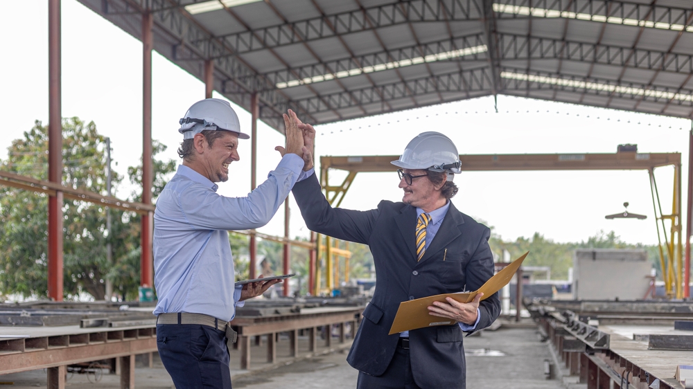 Two men standing outside, in an industrial setting, wearing hard hats, hi-fiving